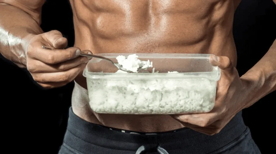 hombre sin camiseta al que se le ven sus abdominales come de un recipiente con una cuchara una comida de arroz
