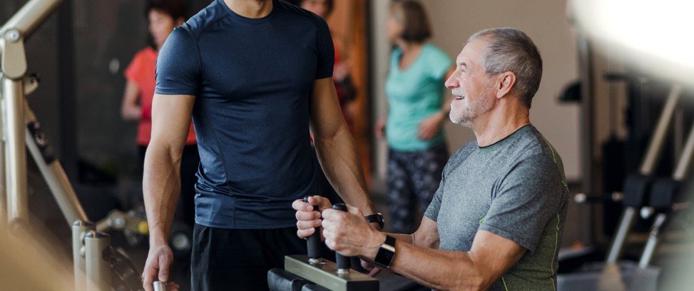 señor mayor escuchando instrucciones de entrenador personal en el gimnasio antes de comenzar a realizar una serie en maquina para fortalecer los brazos