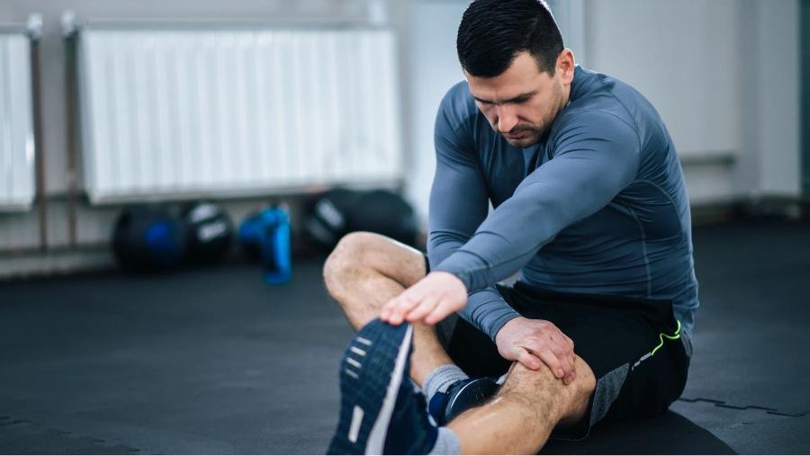 usuario de gimnasio realiza un estiramiento de su pierna derecha mientras permanece sentado en el suelo de un gimnasio