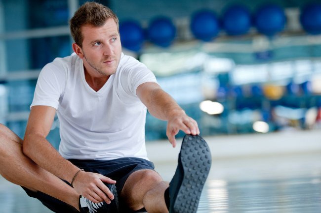 hombre con camiseta blanca y pantalón de deportes negro sentado en el suelo de un gimnasio realiza un estiramiento alcanzando con su mano izquierda su pie izquierdo
